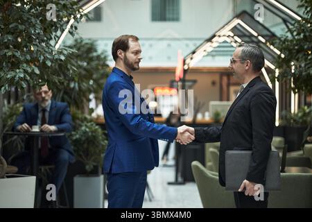 Deux hommes d'affaires serrant la main dans le hall moderne du bureau avec un autre homme assis en arrière-plan. Les deux hommes portent des costumes et sourient, indiquant une réunion d'affaires réussie Banque D'Images