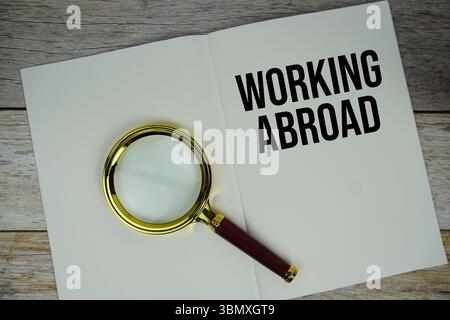 Travailler à l'étranger message texte écrit sur une note de papier avec vue de dessus de loupe sur fond en bois Banque D'Images