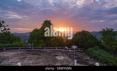 Le soleil se couche derrière les sommets des montagnes au milieu des collines verdoyantes comme le soleil se couche, avec de beaux nuages et la lumière douce du soleil coulant tranquillement vers le bas Banque D'Images