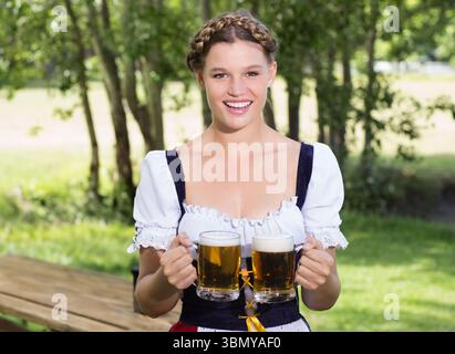 Femme portant la robe traditionnelle dirndl servant deux tasses de bière mousseuse à la table de jardin de bière en plein air Banque D'Images