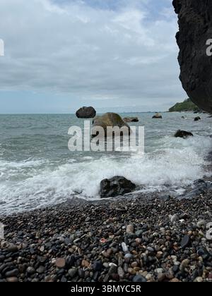 L'image montre une scène côtière avec un premier plan de galets multicolores, des vagues s'écrasant sur le rivage, et plusieurs gros rochers dans la mer. Un sombre Banque D'Images