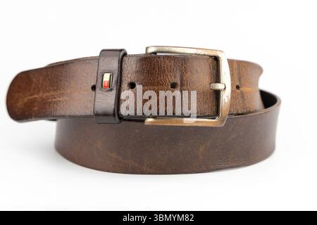 Londres, Royaume-Uni - 29 juin 2025 : ceinture en cuir brun Tommy Hilfiger avec boucle logo et boucle en argent usée roulée soigneusement sur fond blanc. Banque D'Images