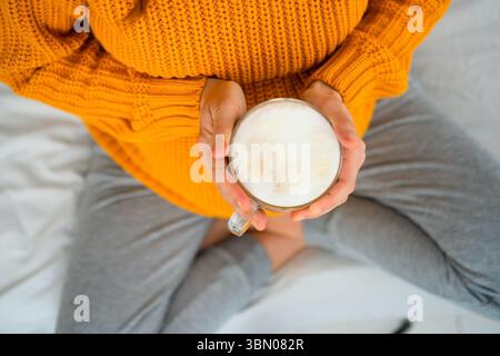 Vue de haut en bas d'une femme enceinte en position de lotus tenant un latte, contemplant les choix alimentaires pendant la grossesse. Moment chaleureux de soins personnels et de conscience. Banque D'Images