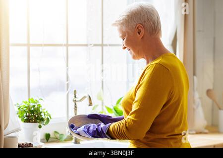 Une femme âgée lave les plats dans la cuisine, en utilisant des gants en caoutchouc Banque D'Images