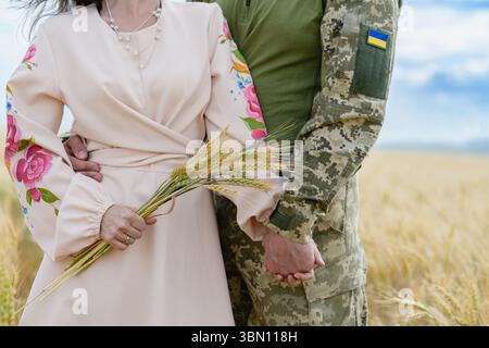 Gros plan d'un soldat ukrainien en uniforme et d'une femme en robe brodée traditionnelle tenant les mains et un bouquet de blé, debout dans un champ mûr sur Banque D'Images