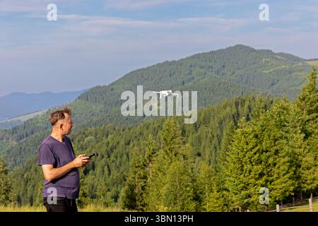 Man Flying Drone au-dessus du paysage forestier de montagne. Personne exploitant un drone avec une télécommande tout en se tenant debout sur une colline herbeuse, surplombant Banque D'Images