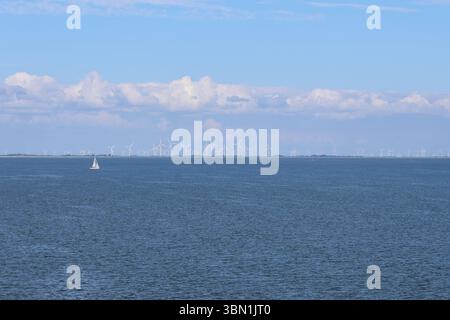 Un voilier solitaire se déplace gracieusement dans des eaux tranquilles, avec un éventail d'éoliennes visibles à l'horizon sous un ciel lumineux et partiellement nuageux. Banque D'Images