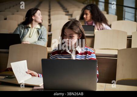 Trois jeunes femmes polyethniques assises dans la salle de conférence de l'université utilisant des ordinateurs portables, une femme caucasienne au premier plan lisant des notes tandis que deux femmes multiethniques en arrière-plan parlent entre elles Banque D'Images