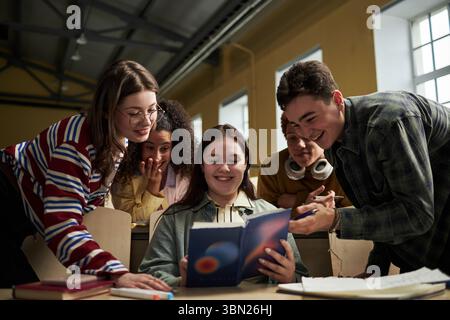 Groupe d'étudiants universitaires multiethniques se réunissant autour du bureau souriant et discutant du manuel ensemble, jeunes adultes collaborant sur un projet académique en salle de classe Banque D'Images
