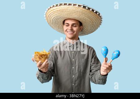 Beau jeune homme en sombrero avec maracas et bol de nachos sur fond bleu Banque D'Images
