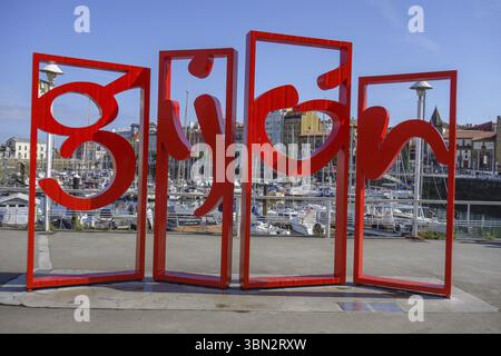 Sculpture en lettres rouges devant un port plein de bateaux sous un ciel ensoleillé, gijon, espagne Banque D'Images