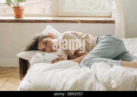 Triste jeune femme regardant son smartphone dans la journée tout en étant allongé sur le lit, la dépendance d'Internet Banque D'Images