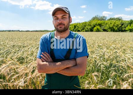 Homme fermier en casquette et t-shirt bleu debout avec les bras croisés près du champ de céréales Banque D'Images