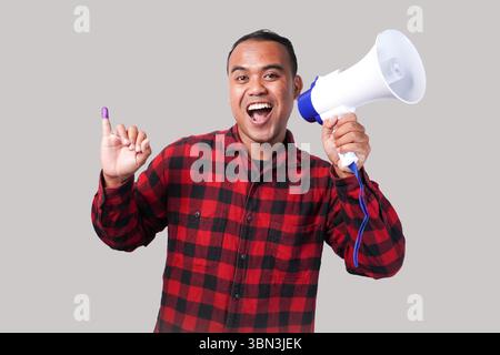 Homme indonésien énergique en tenue de chemise en flanelle , tenant et criant au mégaphone, montrant fièrement petit doigt trempé dans l'encre violette après le vote Banque D'Images