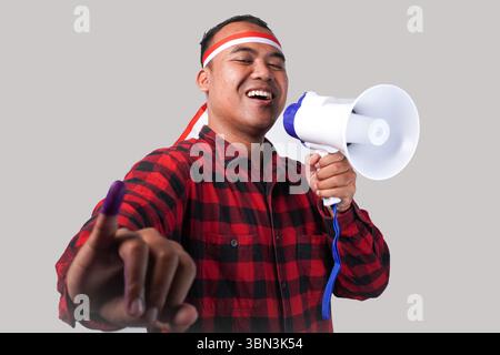 Homme indonésien énergique en tenue de chemise en flanelle, bandeau drapeau , tenant et criant au mégaphone, montrant fièrement petit doigt trempé dans le violet Banque D'Images