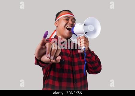 Homme indonésien énergique en tenue de chemise en flanelle, bandeau drapeau , tenant et criant au mégaphone, montrant fièrement petit doigt trempé dans le violet Banque D'Images