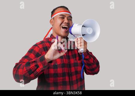 Homme indonésien énergique en tenue de chemise en flanelle, bandeau drapeau , tenant et criant au mégaphone, montrant fièrement petit doigt trempé dans le violet Banque D'Images