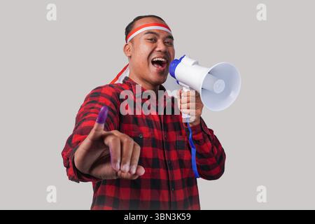 Homme indonésien énergique en tenue de chemise en flanelle, bandeau drapeau , tenant et criant au mégaphone, montrant fièrement petit doigt trempé dans le violet Banque D'Images