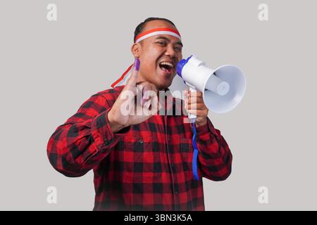 Homme indonésien énergique en tenue de chemise en flanelle, bandeau drapeau , tenant et criant au mégaphone, montrant fièrement petit doigt trempé dans le violet Banque D'Images