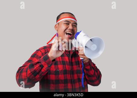 Homme indonésien énergique en tenue de chemise en flanelle, bandeau drapeau , tenant et criant au mégaphone, montrant fièrement petit doigt trempé dans le violet Banque D'Images