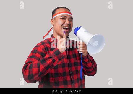 Homme indonésien énergique en tenue de chemise en flanelle, bandeau drapeau , tenant et criant au mégaphone, montrant fièrement petit doigt trempé dans le violet Banque D'Images