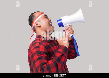 Homme indonésien énergique en tenue de chemise en flanelle, bandeau drapeau , tenant et criant au mégaphone, montrant fièrement petit doigt trempé dans le violet Banque D'Images