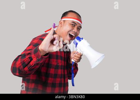 Homme indonésien énergique en tenue de chemise en flanelle, bandeau drapeau , tenant et criant au mégaphone, montrant fièrement petit doigt trempé dans le violet Banque D'Images