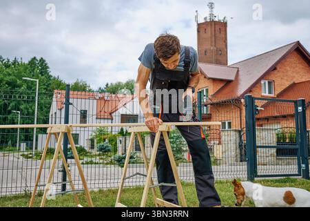 Homme utilisant la scie sauteuse électrique pour couper la planche de bois à l'extérieur près des bâtiments résidentiels. Menuiserie extérieure et concept de travail de bricolage et d'amélioration de la maison Banque D'Images