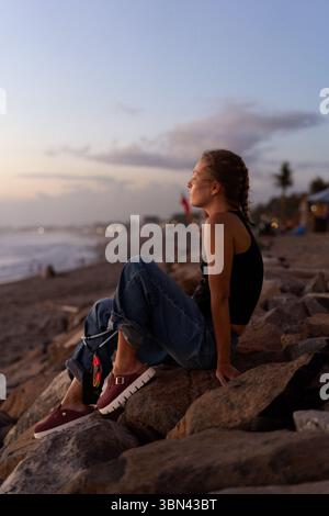 Jeune femme assise sur des rochers au bar de la plage pendant l'heure dorée Banque D'Images