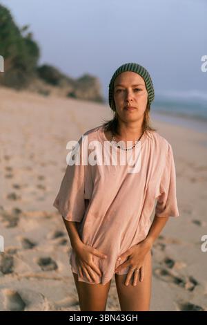 Jeune femme en chemise rose et bonnet vert se tient sur la plage au lever du soleil Banque D'Images