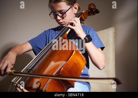 Un jeune individu est vu jouer intensément du violoncelle, se concentrant sur la maîtrise de leur performance. Le cadre intérieur chaleureux souligne leur dévouement et Banque D'Images