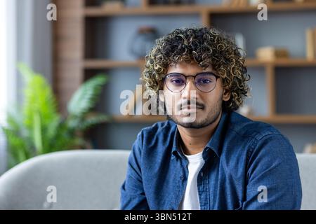 Un jeune homme aux cheveux bouclés et aux lunettes pose à l'intérieur, regardant directement la caméra, avec une expression neutre. Banque D'Images