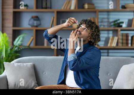 Un homme aux cheveux bouclés applique des gouttes pour les yeux, assis sur un canapé, avec une étagère en arrière-plan. Il semble ressentir une gêne oculaire. Banque D'Images