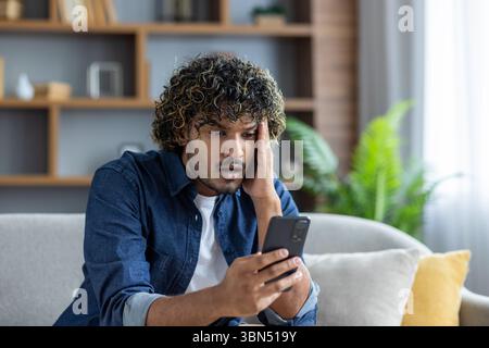 Un jeune homme regarde son téléphone avec une expression inquiète tout en étant assis sur un canapé à la maison. Banque D'Images