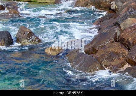 Vue rapprochée des vagues de l'océan s'écrasant contre les rochers côtiers accidentés. L'eau de mer turquoise tourbillonnante et bleu profond crée une pulvérisation blanche spectaculaire Banque D'Images