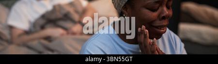 Femme noire d'âge moyen pleurant les mains sur le visage assis au premier plan, tandis que l'homme caucasien d'âge moyen assis dans le lit en arrière-plan regardant loin pendant la situation de conflit Banque D'Images