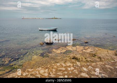 Petit bateau blanc flottant sur la mer calme près de la rive rocheuse. Phare sur l'île de Barbaran en arrière-plan. Scène côtière paisible, eau claire, été t Banque D'Images
