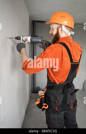 Portrait de perçant concentré barbu ouvrier de construction trou avec perceuse électrique dans le mur. Constructeur professionnel et concept de contremaître Banque D'Images
