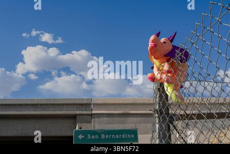 Un dragon en peluche coloré pris sur une clôture à maillons près d'un passage supérieur d'autoroute à Barstow, en Californie, avec un panneau routier pointant vers San Bernardin Banque D'Images