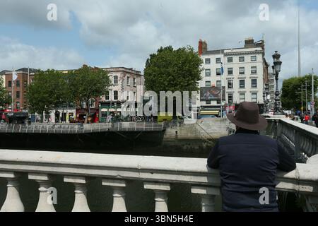 Dublin, Irlande - 4 juin 2025 - Un homme avec un chapeau Fedora regarde de l'autre côté de la rivière Liffey à côté du pont O'Connell dans le centre-ville de Dublin par un après-midi nuageux Banque D'Images