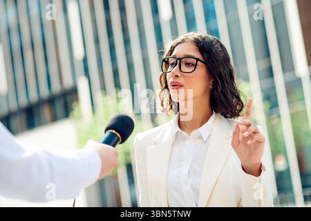 Femme d'affaires confiante interviewée à l'extérieur dans un cadre urbain moderne par une journée ensoleillée, représentant le leadership Banque D'Images