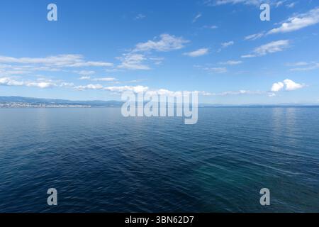 Une mer bleue tranquille s'étend sous un vaste ciel parsemé de nuages blancs, avec un littoral lointain visible à l'horizon. Banque D'Images