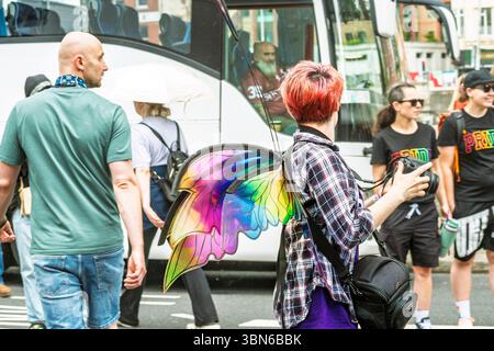 Femme aux cheveux rouges vibrants avec des ailes colorées, caméra à la main, regardant en arrière un vieux chauffeur de bus. Un curieux moment urbain. 28 juin 2025 Banque D'Images