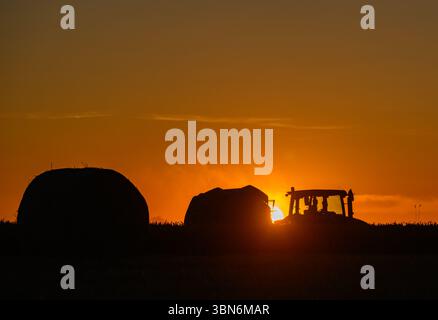Petersdorf, Allemagne. 30 juin 2025. Un agriculteur conduit son tracteur et une presse à paille au-dessus d'un champ de céréales partiellement récolté dans le district d'Oder-Spree dans l'est du Brandebourg tard dans la soirée au coucher du soleil et presse des balles rondes. La récolte des céréales commence actuellement sur les champs en Allemagne. Crédit : Patrick Pleul/dpa/Alamy Live News Banque D'Images
