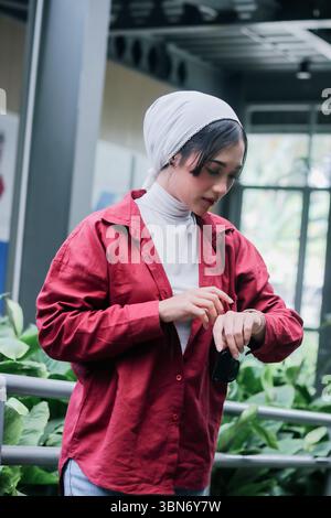 Une jeune femme à la mode portant un foulard blanc et une chemise rouge, regardant attentivement sa smartwatch. Le décor allie design urbain et touche de Banque D'Images