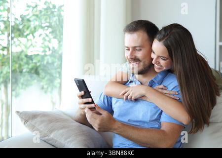 Heureux couple vérifiant le téléphone portable assis sur un canapé à la maison Banque D'Images