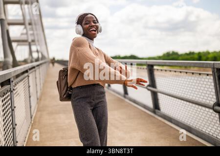 Heureuse jeune femme noire marche sur le chemin de pied de pont, écoute de la musique et sourit. Elle profite d'une journée ensoleillée en plein air. Banque D'Images