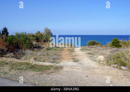 Chypre, Un chemin de terre menant à une vaste étendue d'océan bleu sous un ciel bleu clair et lumineux. La route est flanquée d'un mélange de végétation verte et brune, y compris des buissons et des arbres, et est bordée par une falaise rocheuse basse d'un côté. La perspective est du côté de la route, regardant vers l'océan, créant un sentiment de profondeur et de distance. La route semble être un chemin de terre ou de gravier, et une petite section d'une route pavée est visible dans le coin inférieur gauche. Un œil d'artiste et une imagination façonnée Banque D'Images