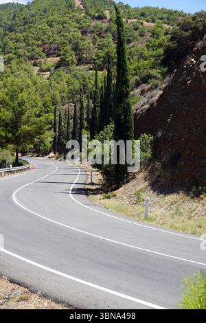 Une route sinueuse qui courbe doucement vers la gauche, entourée d'un paysage verdoyant et luxuriant. La route est bordée par un garde-corps sur le côté gauche et est flanquée de grands cyprès sur le côté droit. La route est pavée d'asphalte et a des marquages de voie blancs. Le terrain environnant est un mélange de végétation verte et de terre brune, avec une falaise rocheuse visible sur le côté droit. Le ciel est d'un bleu clair, et la scène est baignée de lumière naturelle, créant un paysage serein et pittoresque., les sites touristiques de Chypre Banque D'Images