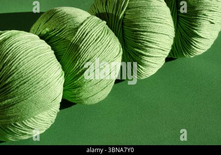 Quatre boules de fil de laine vert clair sur un fond texturé vert foncé. La lumière du soleil de gauche projette des ombres douces, mettant en évidence la texture de la sp Banque D'Images